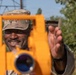 A military service member in camouflage uniform and patrol cap is smiling while operating or adjusting a bright orange surveying instrument, likely a theodolite or transit level. The soldier appears to be conducting land surveying or engineering work in an outdoor environment. The image is taken from the perspective of looking through or past the instrument toward the operator.