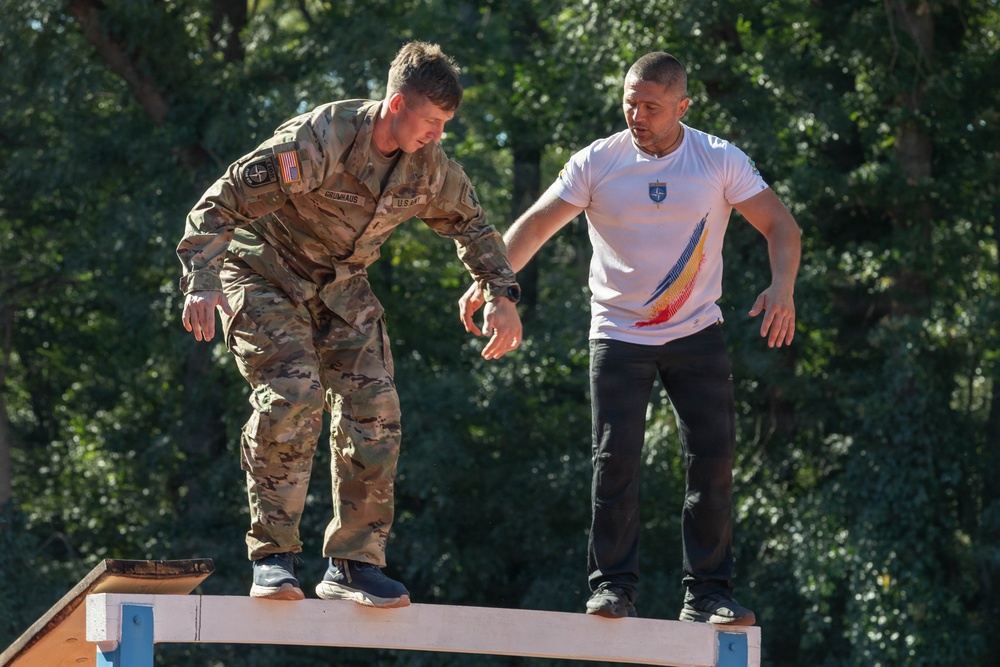 Two individuals are standing on an elevated narrow beam or plank structure, appearing to navigate a balance obstacle course. One person is wearing U.S. Army multicam uniform with an American flag and NATO patch, while the other wears a white t-shirt featuring what appears to be Romanian national colors. The setting appears to be an outdoor military training or obstacle course environment.