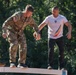 Two individuals are standing on an elevated narrow beam or plank structure, appearing to navigate a balance obstacle course. One person is wearing U.S. Army multicam uniform with an American flag and NATO patch, while the other wears a white t-shirt featuring what appears to be Romanian national colors. The setting appears to be an outdoor military training or obstacle course environment.