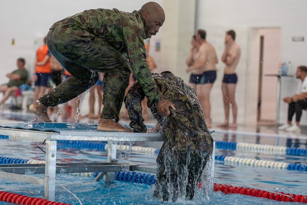 Two individuals wearing wet camouflage military uniforms are engaged in what appears to be a water survival or combat swimming training exercise at an indoor swimming pool facility. One person is crouching on a metal platform at the pool's edge while pulling or assisting another person who is emerging from the water. Several observers in swimwear are visible in the background watching the exercise.