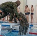 Two individuals wearing wet camouflage military uniforms are engaged in what appears to be a water survival or combat swimming training exercise at an indoor swimming pool facility. One person is crouching on a metal platform at the pool's edge while pulling or assisting another person who is emerging from the water. Several observers in swimwear are visible in the background watching the exercise.