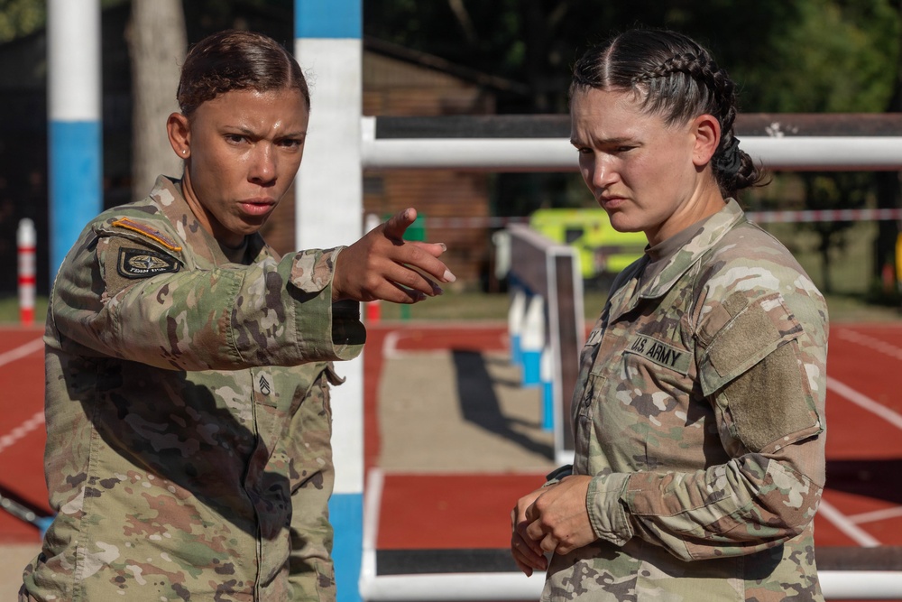 Two female U.S. Army soldiers in MultiCam (OCP) camouflage uniforms are engaged in a focused discussion at what appears to be an outdoor athletic track or training facility. The soldier on the left is pointing and appears to be giving instructions or directions to the soldier on the right. Both display serious, concentrated expressions suggesting a training or competition environment.