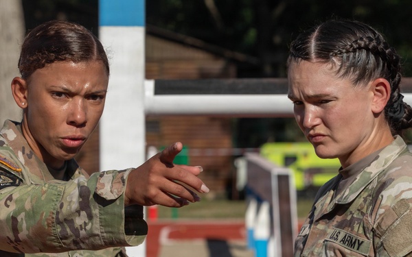 Two female U.S. Army soldiers in MultiCam (OCP) camouflage uniforms are engaged in a focused discussion at what appears to be an outdoor athletic track or training facility. The soldier on the left is pointing and appears to be giving instructions or directions to the soldier on the right. Both display serious, concentrated expressions suggesting a training or competition environment.