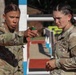 Two female U.S. Army soldiers in MultiCam (OCP) camouflage uniforms are engaged in a focused discussion at what appears to be an outdoor athletic track or training facility. The soldier on the left is pointing and appears to be giving instructions or directions to the soldier on the right. Both display serious, concentrated expressions suggesting a training or competition environment.