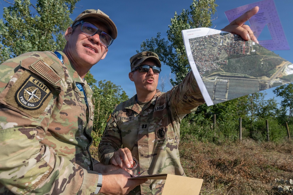 Two U.S. Army soldiers in MultiCam/OCP camouflage uniforms are conducting map reconnaissance outdoors in a vegetated area. One soldier holds up what appears to be a printed aerial/satellite imagery map while the other holds a compass and a clipboard or tablet. They appear to be engaged in land navigation or mission planning activities.