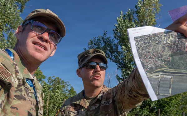 Two U.S. Army soldiers in MultiCam/OCP camouflage uniforms are conducting map reconnaissance outdoors in a vegetated area. One soldier holds up what appears to be a printed aerial/satellite imagery map while the other holds a compass and a clipboard or tablet. They appear to be engaged in land navigation or mission planning activities.