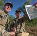 Two U.S. Army soldiers in MultiCam/OCP camouflage uniforms are conducting map reconnaissance outdoors in a vegetated area. One soldier holds up what appears to be a printed aerial/satellite imagery map while the other holds a compass and a clipboard or tablet. They appear to be engaged in land navigation or mission planning activities.