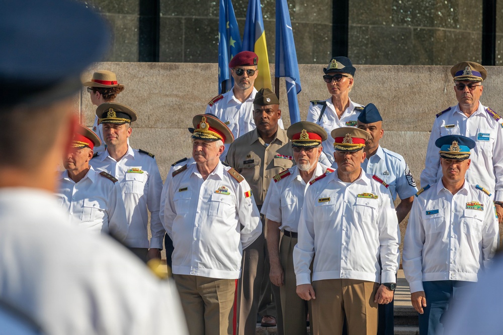 A formal military ceremony featuring personnel in various national uniforms gathered in a group formation. Multiple flags are visible in the background, including what appears to be the EU flag, NATO flag, and a yellow-and-blue flag. The gathering appears to be a multinational military or diplomatic event.