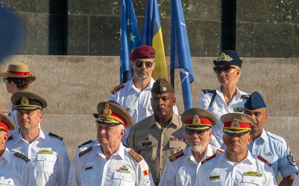 A formal military ceremony featuring personnel in various national uniforms gathered in a group formation. Multiple flags are visible in the background, including what appears to be the EU flag, NATO flag, and a yellow-and-blue flag. The gathering appears to be a multinational military or diplomatic event.