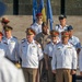 A formal military ceremony featuring personnel in various national uniforms gathered in a group formation. Multiple flags are visible in the background, including what appears to be the EU flag, NATO flag, and a yellow-and-blue flag. The gathering appears to be a multinational military or diplomatic event.