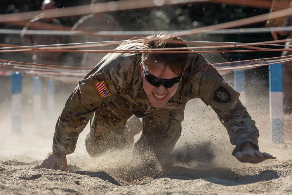 A U.S. Army soldier in MultiCam (OCP) camouflage uniform is crawling under wire obstacles through sandy terrain during what appears to be a military obstacle course or training exercise. The soldier is wearing sunglasses and has a U.S. flag patch visible on the right shoulder, along with additional unit patches. Sand is visibly displaced around the soldier's hands and body, indicating rapid, forceful movement.