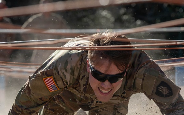 A U.S. Army soldier in MultiCam (OCP) camouflage uniform is crawling under wire obstacles through sandy terrain during what appears to be a military obstacle course or training exercise. The soldier is wearing sunglasses and has a U.S. flag patch visible on the right shoulder, along with additional unit patches. Sand is visibly displaced around the soldier's hands and body, indicating rapid, forceful movement.