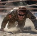 A U.S. Army soldier in MultiCam (OCP) camouflage uniform is crawling under wire obstacles through sandy terrain during what appears to be a military obstacle course or training exercise. The soldier is wearing sunglasses and has a U.S. flag patch visible on the right shoulder, along with additional unit patches. Sand is visibly displaced around the soldier's hands and body, indicating rapid, forceful movement.