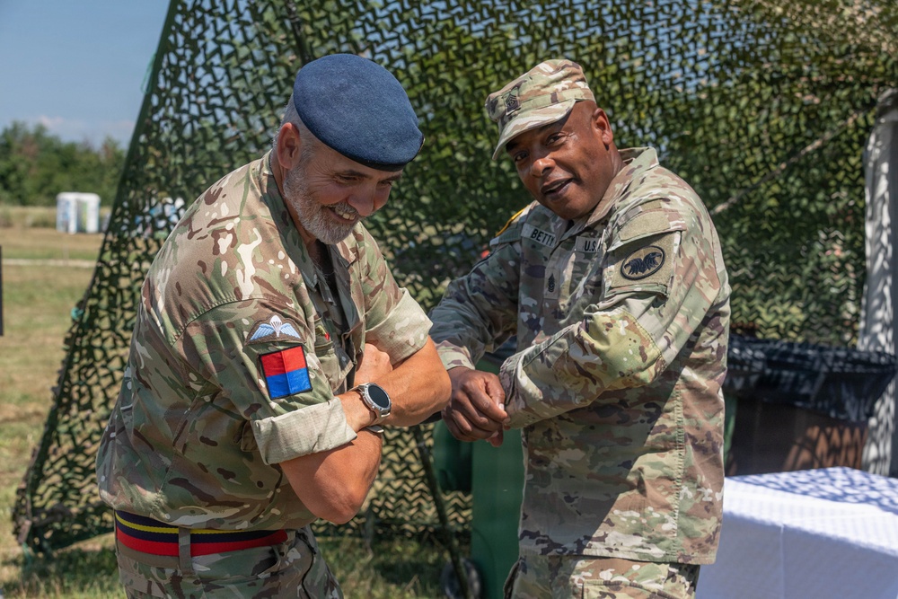 Two military personnel in camouflage uniforms are engaged in what appears to be a coin or item exchange ceremony outdoors. One soldier wears a British Army uniform with a blue beret and unit patches, while the other wears a U.S. Army uniform with a patrol cap. They are standing in front of a camouflage netting backdrop, both smiling.