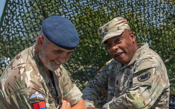 Two military personnel in camouflage uniforms are engaged in what appears to be a coin or item exchange ceremony outdoors. One soldier wears a British Army uniform with a blue beret and unit patches, while the other wears a U.S. Army uniform with a patrol cap. They are standing in front of a camouflage netting backdrop, both smiling.