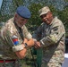 Two military personnel in camouflage uniforms are engaged in what appears to be a coin or item exchange ceremony outdoors. One soldier wears a British Army uniform with a blue beret and unit patches, while the other wears a U.S. Army uniform with a patrol cap. They are standing in front of a camouflage netting backdrop, both smiling.
