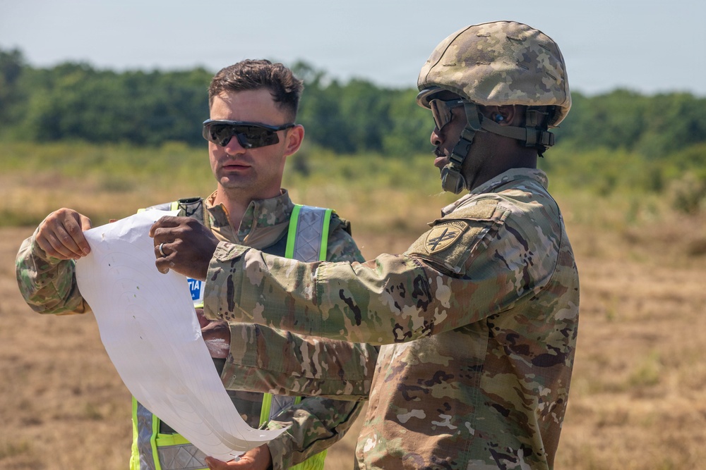 Two military personnel in U.S. Army OCP (Operational Camouflage Pattern) uniforms are reviewing a document or map in an open field environment. One soldier is wearing a combat helmet and pointing at the document, while the other wears sunglasses and a high-visibility safety vest over his uniform. The setting appears to be a training area or operational field site.