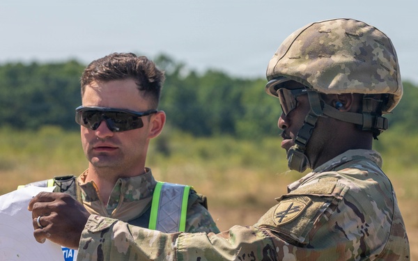 Two military personnel in U.S. Army OCP (Operational Camouflage Pattern) uniforms are reviewing a document or map in an open field environment. One soldier is wearing a combat helmet and pointing at the document, while the other wears sunglasses and a high-visibility safety vest over his uniform. The setting appears to be a training area or operational field site.