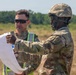 Two military personnel in U.S. Army OCP (Operational Camouflage Pattern) uniforms are reviewing a document or map in an open field environment. One soldier is wearing a combat helmet and pointing at the document, while the other wears sunglasses and a high-visibility safety vest over his uniform. The setting appears to be a training area or operational field site.