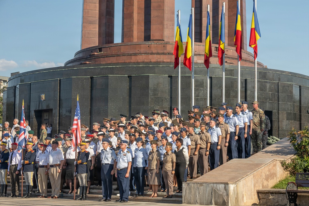 A multinational military ceremony is taking place in front of a large circular monument building, likely in Romania based on the Romanian tricolor flags (blue, yellow, red) prominently displayed. Numerous military personnel in various national uniforms are assembled in formation, with flag bearers carrying the American flag and what appears to be a British flag visible in the foreground. The event appears to be a formal parade or commemorative ceremony at what resembles the Carol Park Mausoleum or a similar Romanian national monument.
