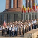 A multinational military ceremony is taking place in front of a large circular monument building, likely in Romania based on the Romanian tricolor flags (blue, yellow, red) prominently displayed. Numerous military personnel in various national uniforms are assembled in formation, with flag bearers carrying the American flag and what appears to be a British flag visible in the foreground. The event appears to be a formal parade or commemorative ceremony at what resembles the Carol Park Mausoleum or a similar Romanian national monument.