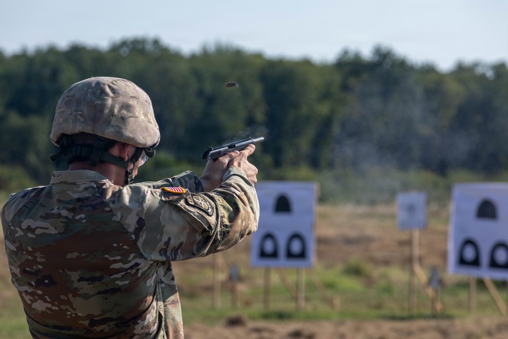 A U.S. Army soldier in MultiCam (OCP) camouflage uniform and combat helmet is firing a semi-automatic pistol at a military shooting range. A spent brass casing is visibly ejecting from the firearm, and smoke is present near the targets downrange. Multiple standard silhouette paper targets are positioned at varying distances on the range.