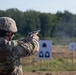 A U.S. Army soldier in MultiCam (OCP) camouflage uniform and combat helmet is firing a semi-automatic pistol at a military shooting range. A spent brass casing is visibly ejecting from the firearm, and smoke is present near the targets downrange. Multiple standard silhouette paper targets are positioned at varying distances on the range.