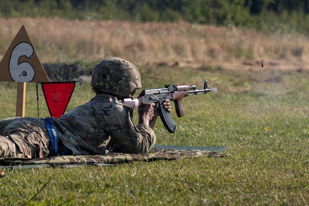 A soldier in camouflage uniform and helmet is firing an AK-pattern rifle from a prone position on a shooting range. A spent brass casing is visibly ejecting from the weapon mid-fire. The shooter is positioned at lane number 6, marked by a wooden sign with a red triangle indicator.