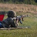 A soldier in camouflage uniform and helmet is firing an AK-pattern rifle from a prone position on a shooting range. A spent brass casing is visibly ejecting from the weapon mid-fire. The shooter is positioned at lane number 6, marked by a wooden sign with a red triangle indicator.