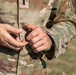 A person wearing a MultiCam/OCP camouflage military uniform is handling what appears to be a firearm magazine and a single round of ammunition. The image is a close-up torso shot focusing on the hands and uniform. The setting appears to be an outdoor training or range environment.