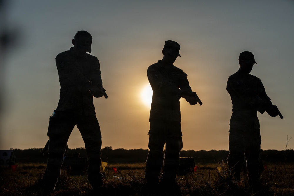 Three silhouetted individuals in military-style uniforms are photographed against a sunset sky in an open field. Each person is holding what appears to be a handgun, suggesting a firearms training or qualification exercise. The backlit composition creates strong silhouettes with limited detail visibility.