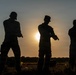 Three silhouetted individuals in military-style uniforms are photographed against a sunset sky in an open field. Each person is holding what appears to be a handgun, suggesting a firearms training or qualification exercise. The backlit composition creates strong silhouettes with limited detail visibility.
