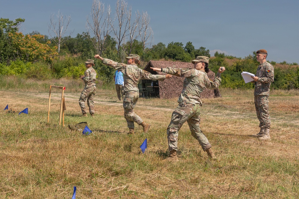 Military personnel in camouflage uniforms are conducting what appears to be a combatives or hand-to-hand combat training exercise in an open field. Two soldiers in the foreground are engaged in a sparring or technique demonstration while a third soldier wearing a brown beret observes and takes notes. Additional soldiers and a camouflage-netted structure are visible in the background.
