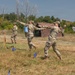 Military personnel in camouflage uniforms are conducting what appears to be a combatives or hand-to-hand combat training exercise in an open field. Two soldiers in the foreground are engaged in a sparring or technique demonstration while a third soldier wearing a brown beret observes and takes notes. Additional soldiers and a camouflage-netted structure are visible in the background.