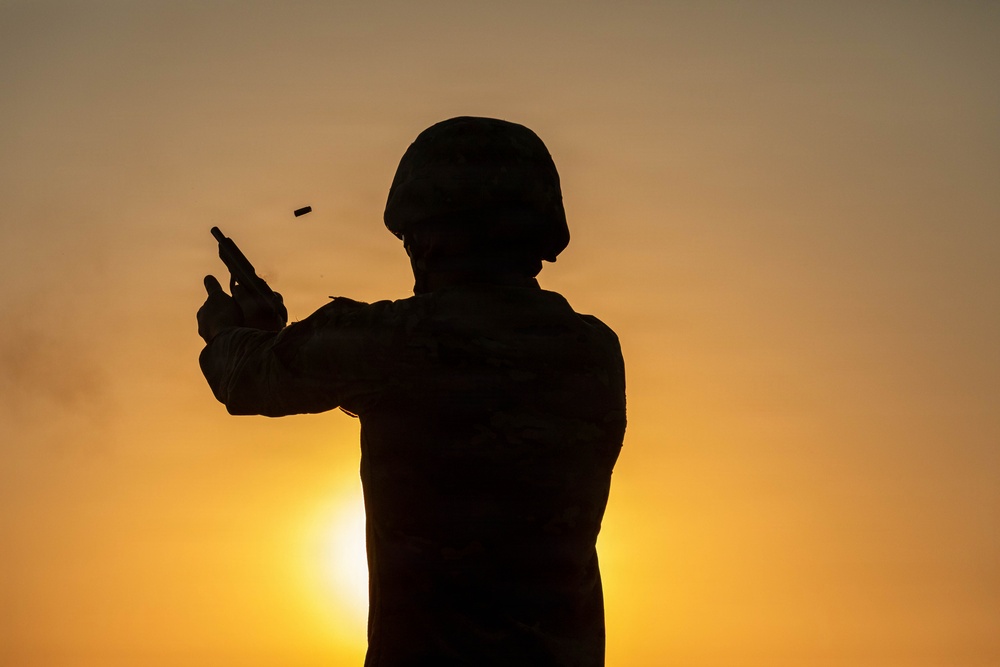 A silhouetted figure wearing a military helmet and tactical gear is firing a handgun against a warm sunset sky. A spent brass casing is visibly ejecting from the firearm, captured mid-air to the left of the weapon. The backlit composition creates a dramatic contrast between the dark figure and the orange-golden sky.