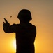 A silhouetted figure wearing a military helmet and tactical gear is firing a handgun against a warm sunset sky. A spent brass casing is visibly ejecting from the firearm, captured mid-air to the left of the weapon. The backlit composition creates a dramatic contrast between the dark figure and the orange-golden sky.