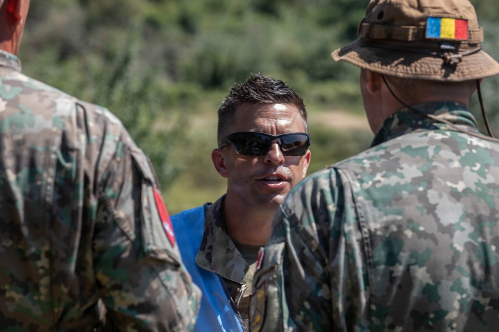 Three military personnel are engaged in conversation outdoors. The central figure wears sunglasses and a blue vest over camouflage uniform, while the figure on the right wears a boonie hat bearing what appears to be the Romanian flag patch. The setting appears to be a field or training environment with vegetation visible in the background.