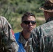 Three military personnel are engaged in conversation outdoors. The central figure wears sunglasses and a blue vest over camouflage uniform, while the figure on the right wears a boonie hat bearing what appears to be the Romanian flag patch. The setting appears to be a field or training environment with vegetation visible in the background.