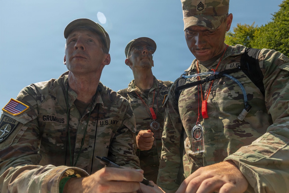 Three U.S. Army soldiers in OCP (Operational Camouflage Pattern) uniforms are engaged in what appears to be a military competition or field exercise. The soldiers show signs of physical exertion with face camouflage paint and are wearing medals/coins on lanyards around their necks. The leftmost soldier, identified by nametape as 'GRUMHAUS,' appears to be writing or reviewing something while the others observe.