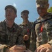 Three U.S. Army soldiers in OCP (Operational Camouflage Pattern) uniforms are engaged in what appears to be a military competition or field exercise. The soldiers show signs of physical exertion with face camouflage paint and are wearing medals/coins on lanyards around their necks. The leftmost soldier, identified by nametape as 'GRUMHAUS,' appears to be writing or reviewing something while the others observe.