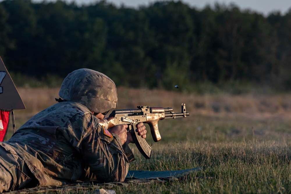 A soldier in US Army camouflage uniform and helmet is firing an AK-pattern rifle from a prone position on a grass field. The image appears to be taken during a training exercise at a shooting range, with a spent casing visibly ejecting from the weapon. The background shows a treeline illuminated by low-angle golden hour lighting.