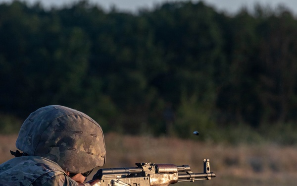 A soldier in US Army camouflage uniform and helmet is firing an AK-pattern rifle from a prone position on a grass field. The image appears to be taken during a training exercise at a shooting range, with a spent casing visibly ejecting from the weapon. The background shows a treeline illuminated by low-angle golden hour lighting.