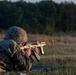 A soldier in US Army camouflage uniform and helmet is firing an AK-pattern rifle from a prone position on a grass field. The image appears to be taken during a training exercise at a shooting range, with a spent casing visibly ejecting from the weapon. The background shows a treeline illuminated by low-angle golden hour lighting.