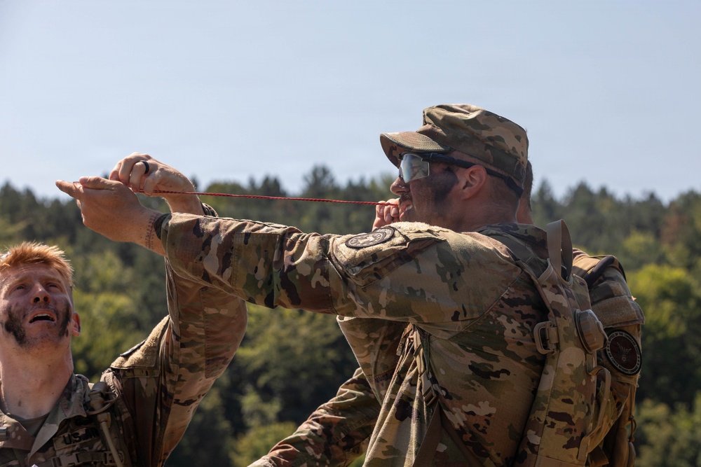 Two military personnel in MultiCam/OCP camouflage uniforms are engaged in an outdoor field activity. The primary subject is pulling or handling a red cord/rope with both hands extended forward, while a second soldier with red hair observes from the left. The setting appears to be a training exercise in a wooded area under clear skies.