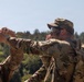 Two military personnel in MultiCam/OCP camouflage uniforms are engaged in an outdoor field activity. The primary subject is pulling or handling a red cord/rope with both hands extended forward, while a second soldier with red hair observes from the left. The setting appears to be a training exercise in a wooded area under clear skies.