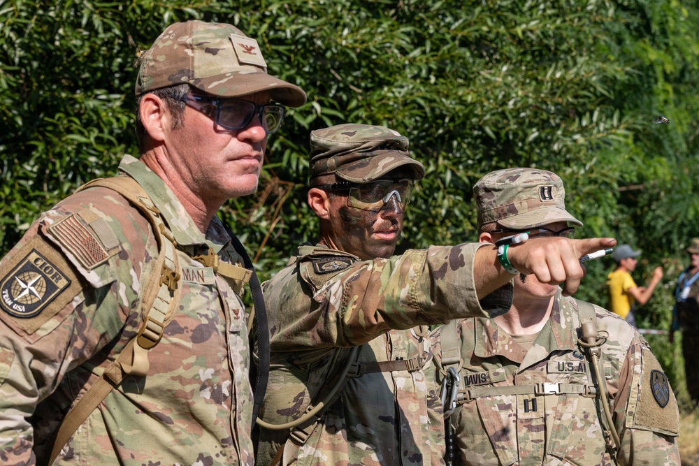 Three U.S. Army soldiers in Operational Camouflage Pattern (OCP) uniforms are gathered outdoors, with one soldier pointing in a specific direction, likely during a training exercise or field operation. Name tapes visible include 'DAVIS' and partially 'MAN', and rank insignia suggest at least one Captain (two vertical bars) is present. A small group of civilians is visible in the background near tree line.