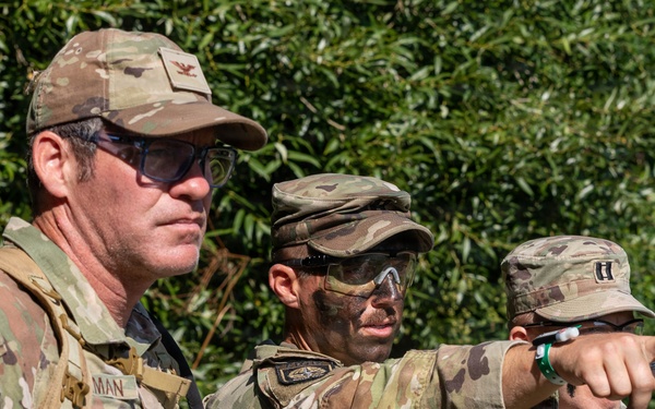 Three U.S. Army soldiers in Operational Camouflage Pattern (OCP) uniforms are gathered outdoors, with one soldier pointing in a specific direction, likely during a training exercise or field operation. Name tapes visible include 'DAVIS' and partially 'MAN', and rank insignia suggest at least one Captain (two vertical bars) is present. A small group of civilians is visible in the background near tree line.