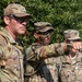 Three U.S. Army soldiers in Operational Camouflage Pattern (OCP) uniforms are gathered outdoors, with one soldier pointing in a specific direction, likely during a training exercise or field operation. Name tapes visible include 'DAVIS' and partially 'MAN', and rank insignia suggest at least one Captain (two vertical bars) is present. A small group of civilians is visible in the background near tree line.