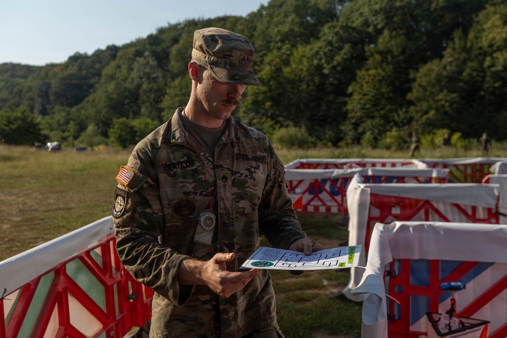 A U.S. Army soldier in OCP (Operational Camouflage Pattern) uniform is reviewing a document or map while standing among red-and-white barrier structures in an outdoor field setting. The soldier wears a name tape, rank insignia, and a U.S. flag patch on the left shoulder, indicating active duty status. The setting appears to be a training exercise or operational planning area with wooded hills in the background.