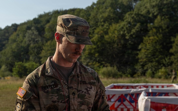 A U.S. Army soldier in OCP (Operational Camouflage Pattern) uniform is reviewing a document or map while standing among red-and-white barrier structures in an outdoor field setting. The soldier wears a name tape, rank insignia, and a U.S. flag patch on the left shoulder, indicating active duty status. The setting appears to be a training exercise or operational planning area with wooded hills in the background.
